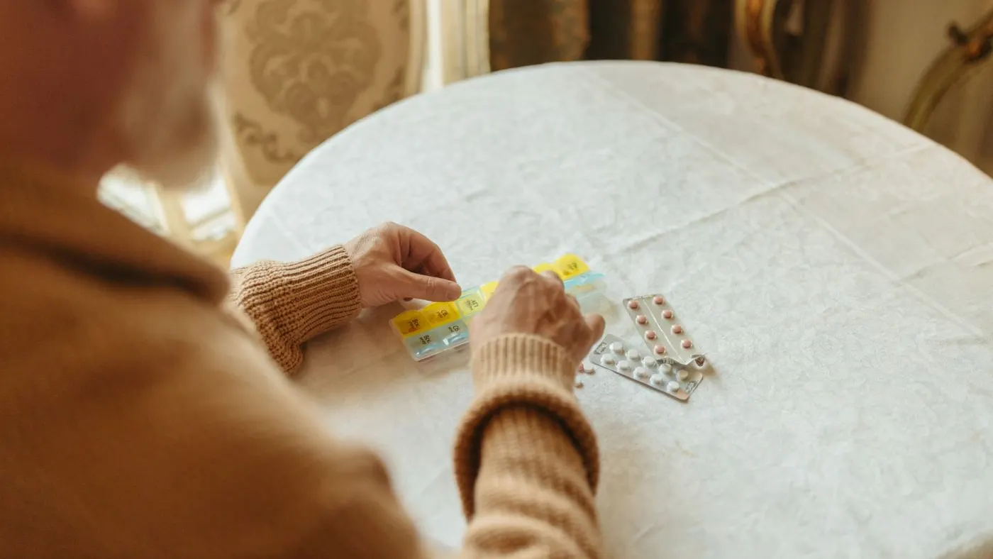 Persona mayor con suéter de lana organizando sus pastillas en un pastillero semanal sobre una mesa con mantel blanco