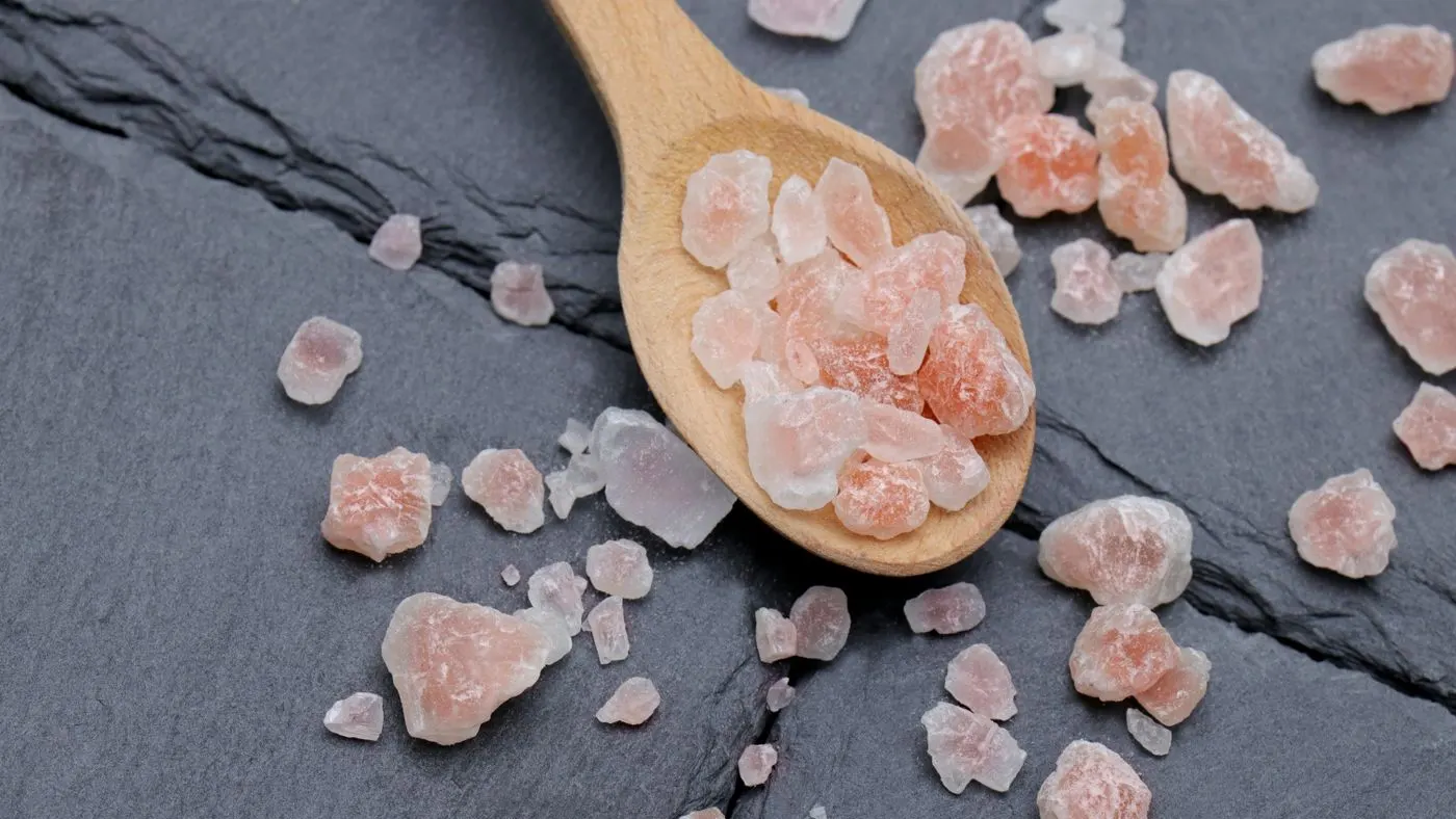 Cuchara de madera con cristales de sal rosada y más cristales regados sobre una superficie de piedra gris oscura