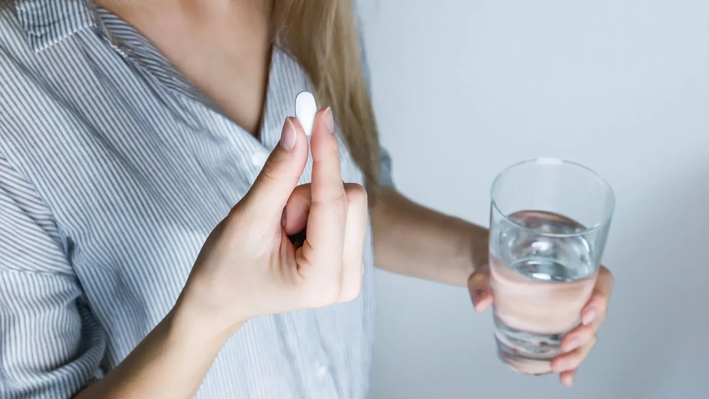 Mujer con camisa de rayas que sostiene una pastilla blanca en una mano y un vaso de agua en la otra, imagen para la guía del valsartán